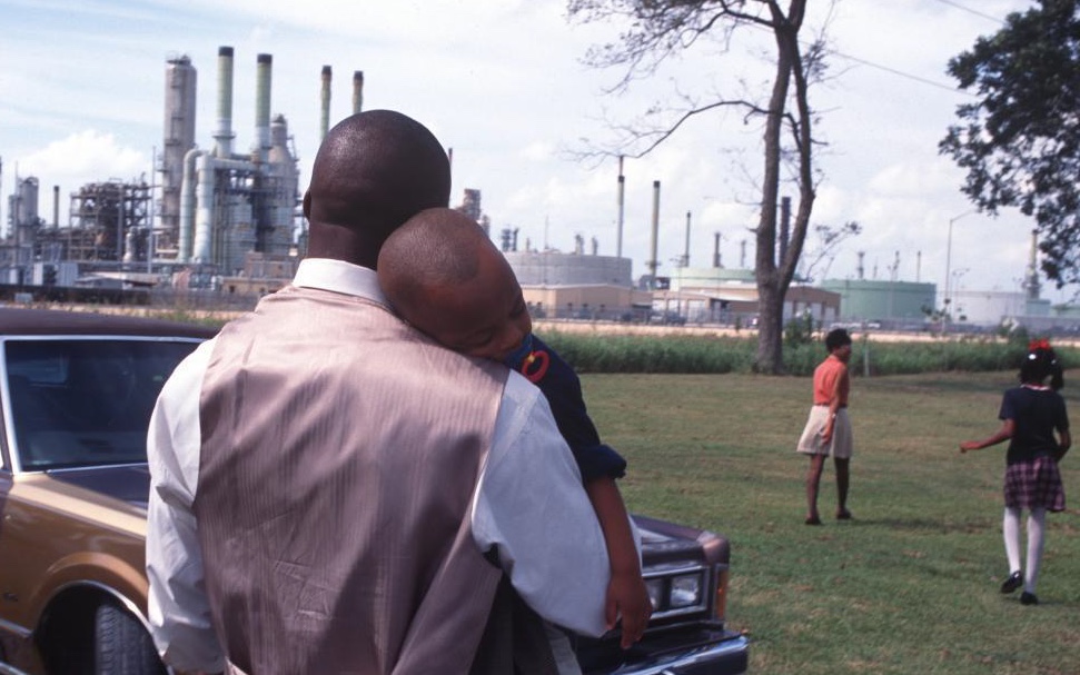 A father holds his child while two women stand nearby in a park beside an industrial facility